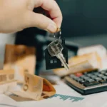 Main tenant des clés de logement avec calculatrice et billets en arrière-plan, illustrant la rentabilité d'un investissement locatif.