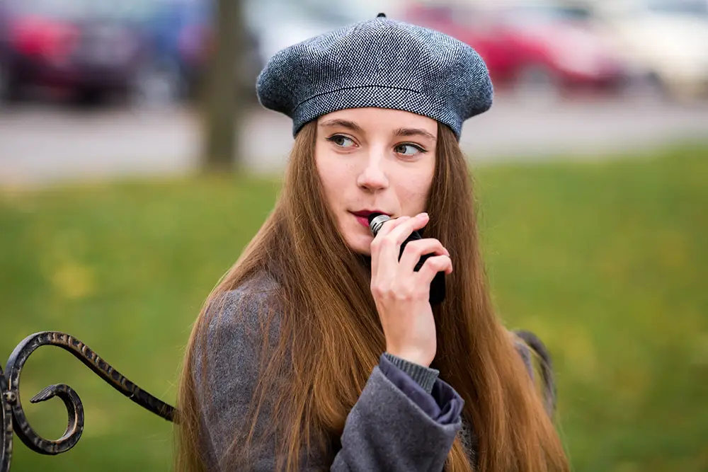 Jeune femme stylée, avec un béret dans un parc en train de vapoter sa cigarette électronique