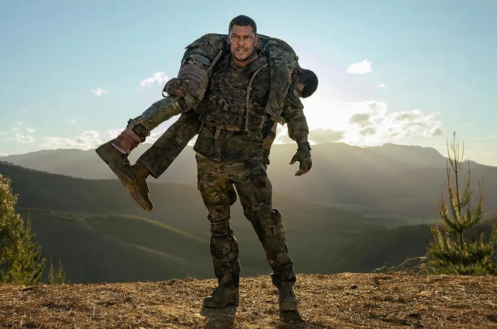 Alan Ritchson debout en haut d'une montagne en portant un soldat blessé